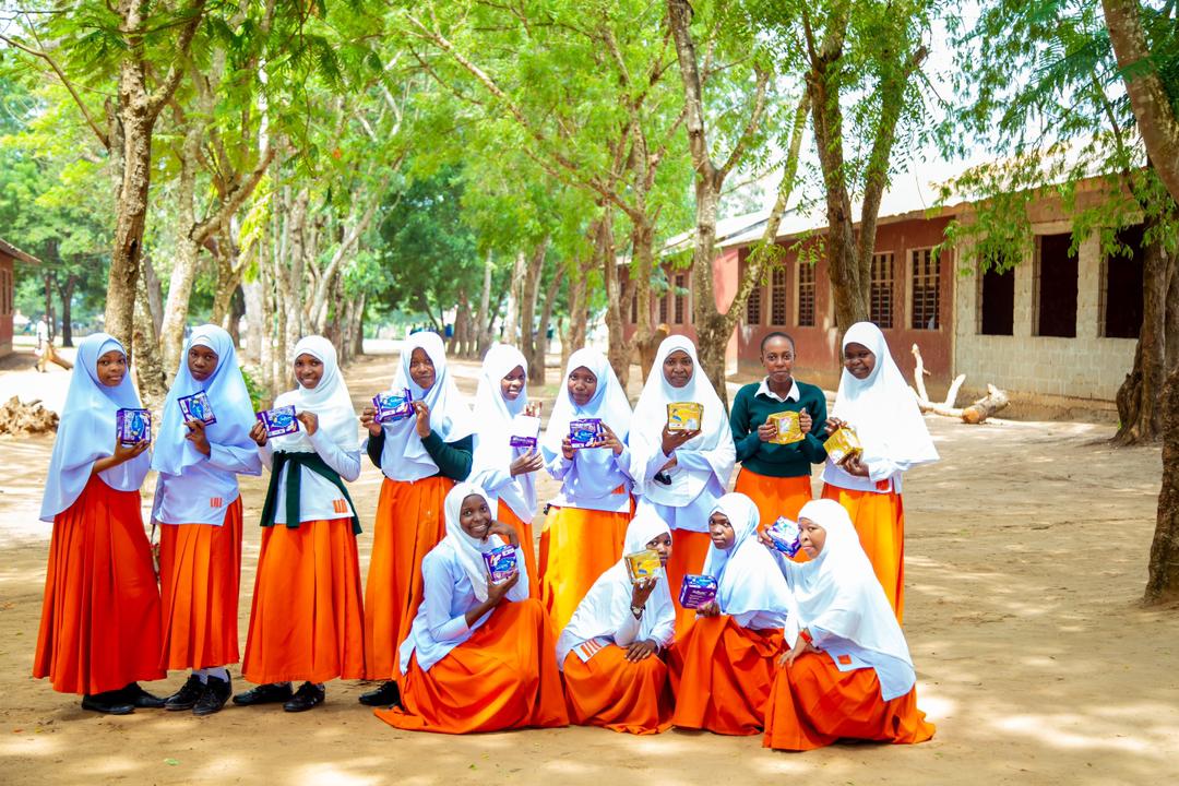 Schoolgirls in orange uniforms standing under trees, each holding a sanitary care package