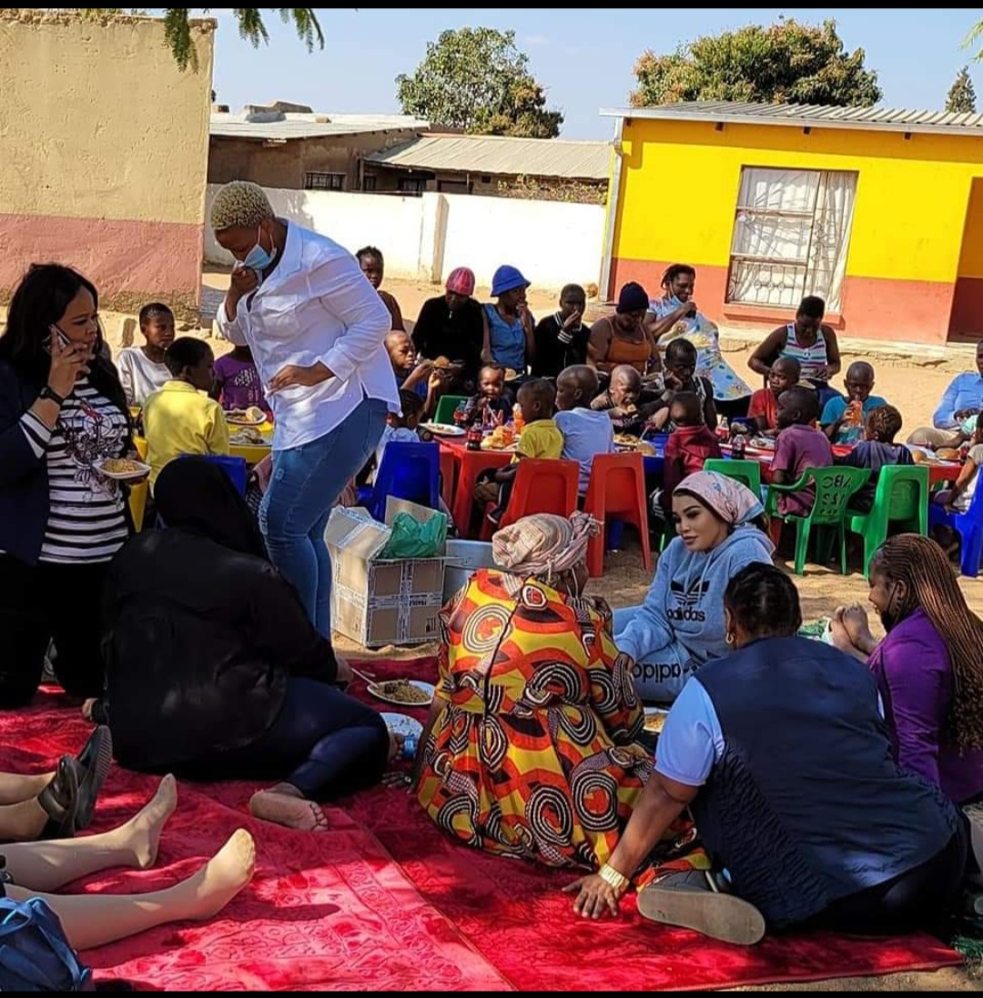 A community meal outdoors with children seated at colourful plastic tables and elders sharing food on mats
