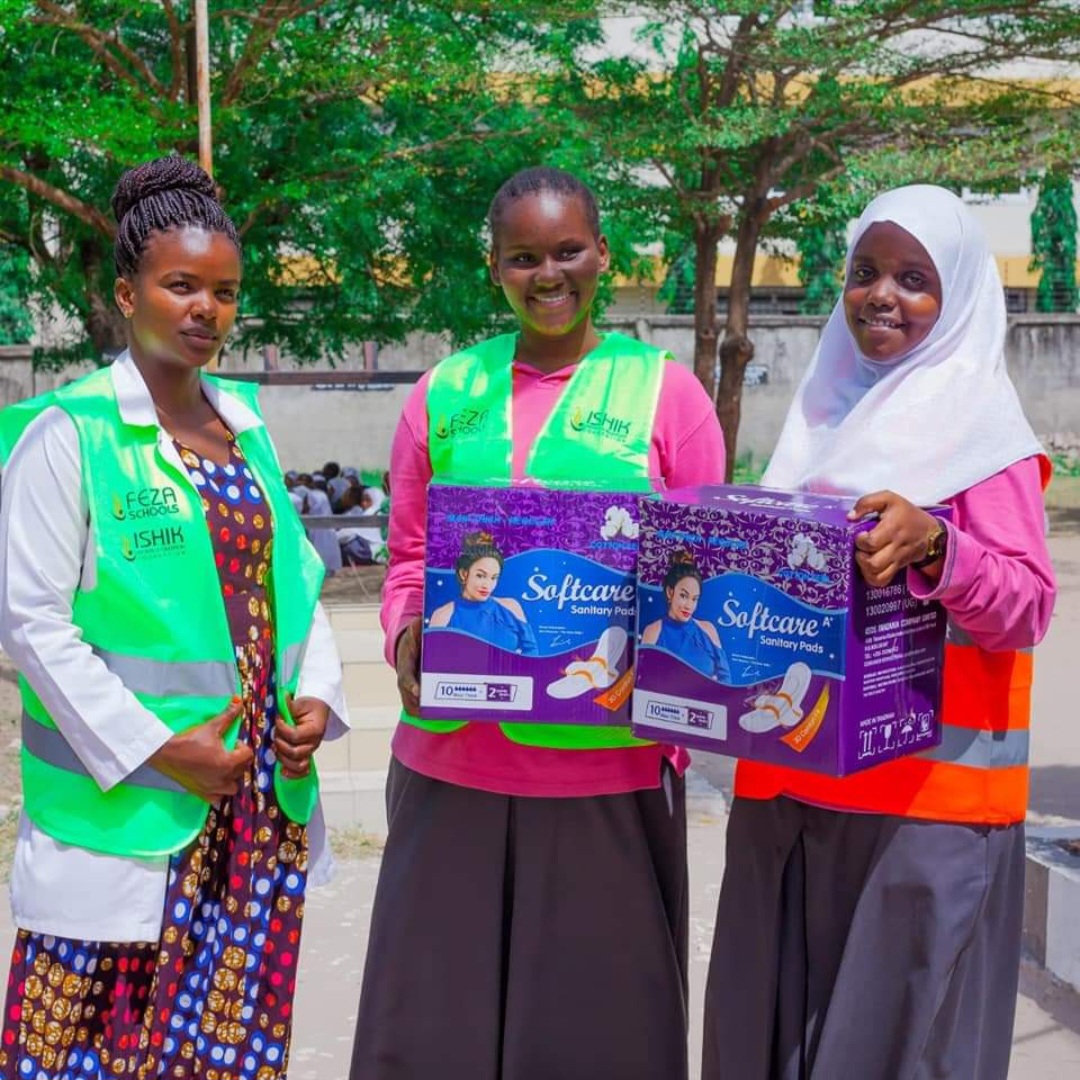 Three schoolgirls in high visibility vests holding Softcare sanitary pad boxes at a school